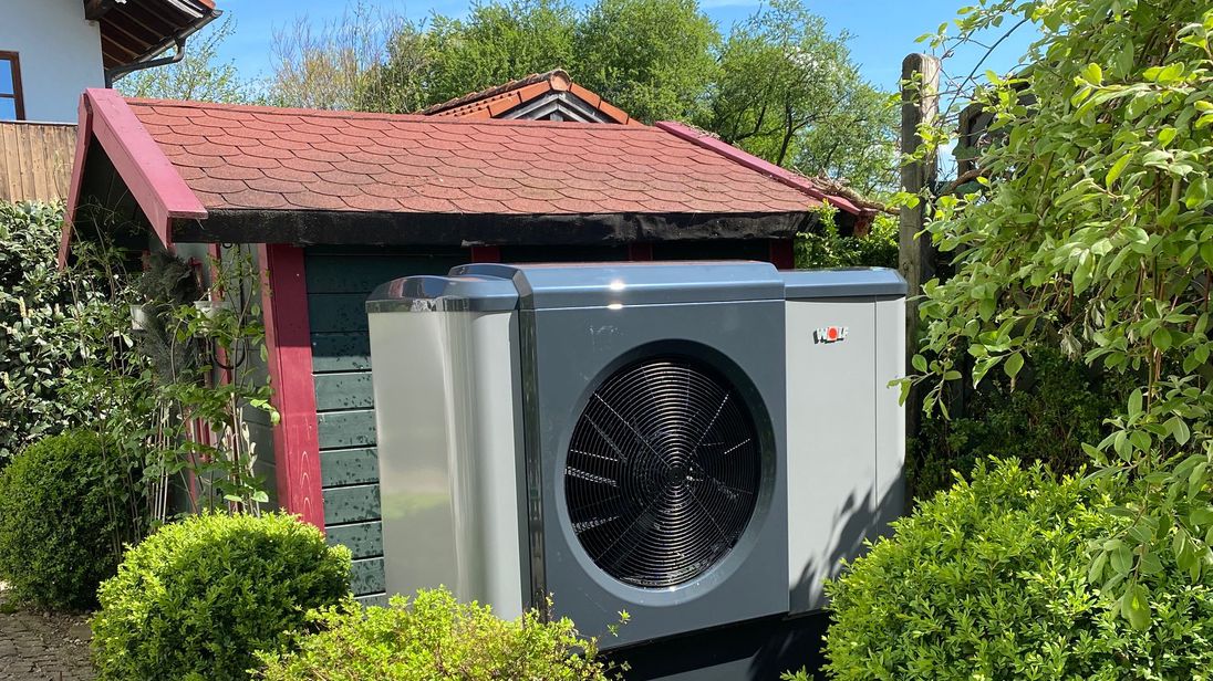 Eine Wärmepumpe in einem Garten vor einem Gartenhaus.